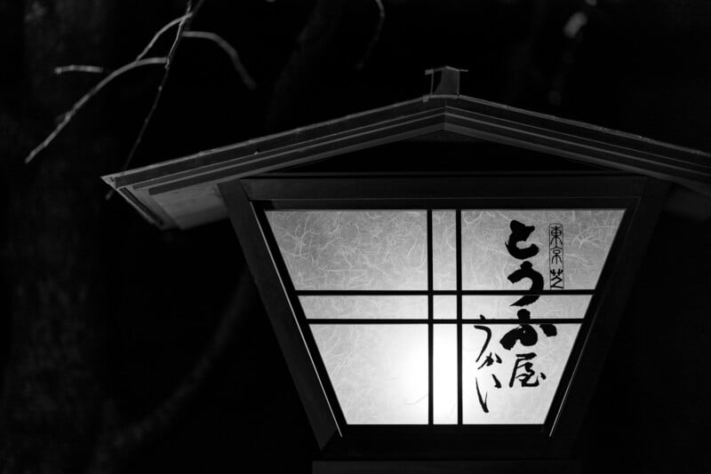 A traditional Japanese lantern glows softly at night, featuring Japanese calligraphy on frosted glass panels. The background is dark, with faint tree branches visible.