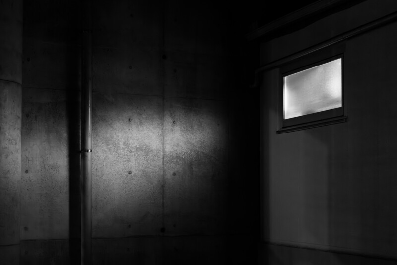 A dimly lit concrete wall with a metal pipe and a small frosted window emitting light on the right side, creating a stark contrast of shadows and illumination.