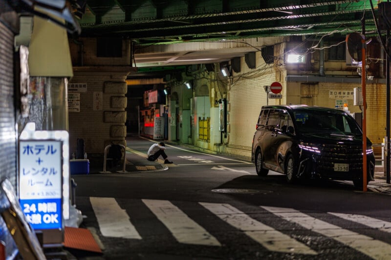 A dimly lit urban street at night under a bridge, with a person crouching near the sidewalk and a black car parked nearby. Japanese signs and storefronts are visible, giving a quiet, late-night city atmosphere.