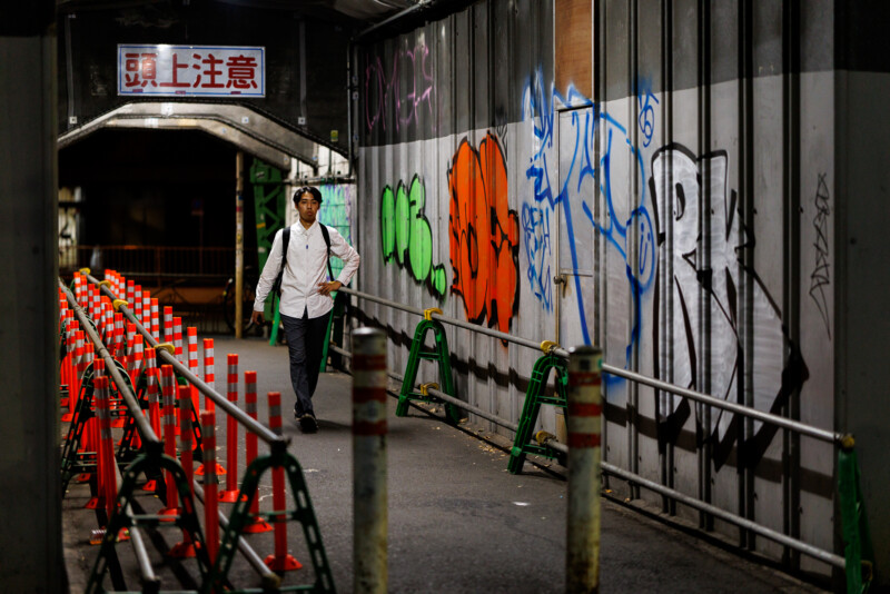 A man in a white shirt and backpack walks through a dimly lit urban passageway lined with orange traffic cones and graffiti-covered walls at night. A sign with Japanese characters hangs overhead.