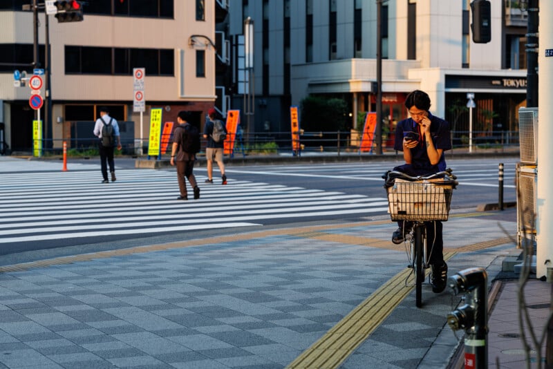 A person rides a bicycle on a city sidewalk, looking at their phone. The bike has a basket in front. In the background, people cross at a wide crosswalk with buildings and street signs visible.