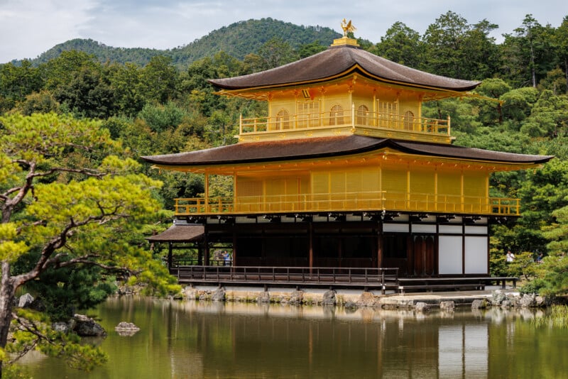 A two-story golden pavilion stands beside a calm pond, surrounded by lush green trees and distant hills under a partly cloudy sky.