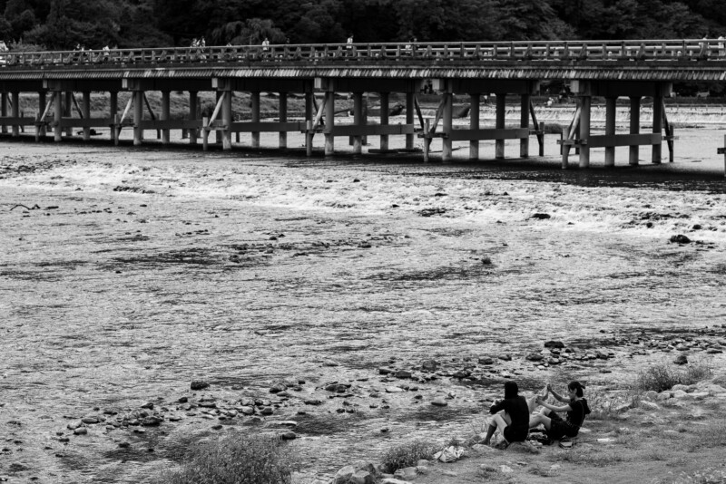 Two people sit on the rocky riverbank in front of a wide wooden bridge spanning a flowing river, with more people walking across the bridge in the background. The image is in black and white.
