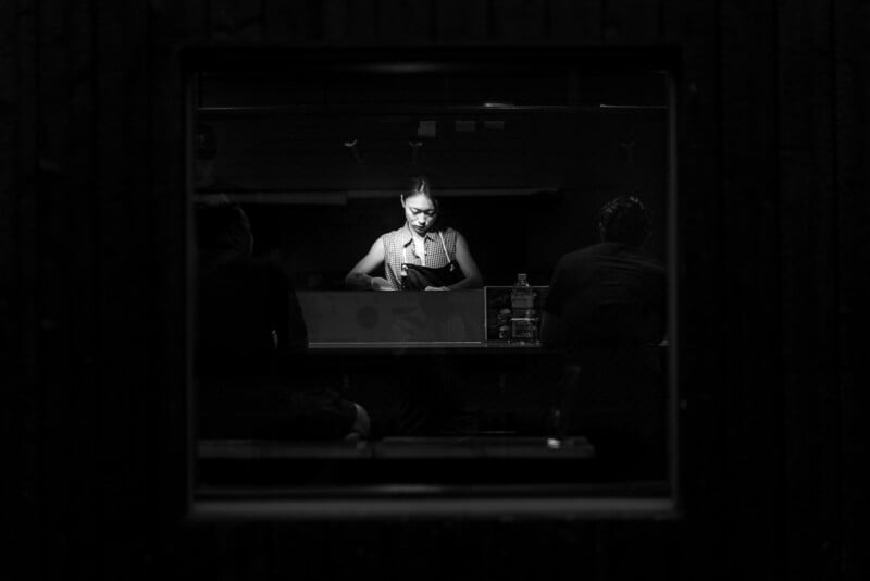 A woman illuminated by a spotlight stands behind a counter, holding a notebook, with two people sitting in shadow on either side. The scene is framed by a window in a dark wall. The image is in black and white.
