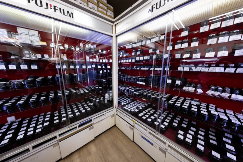 Glass display cases filled with neatly arranged camera lenses and equipment, labeled with price tags, in a store with Fujifilm branding on the top panels. The floor is light wood.