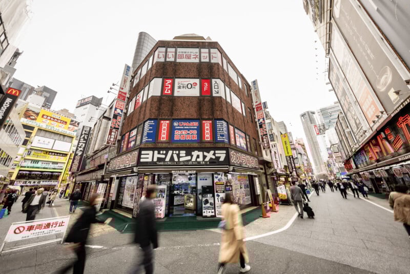 A busy street scene in Japan with people walking past a large electronics store called Yodobashi Camera, surrounded by tall buildings covered in colorful signs and advertisements.