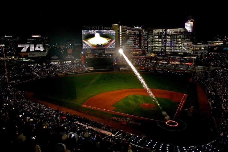 A night view of a brightly lit baseball stadium with a firework shooting up from the field and a large crowd in the stands. The scoreboard and city lights are visible in the background.
