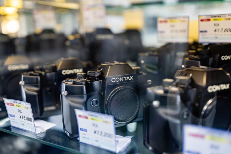 Several black Contax film cameras are displayed on glass shelves, each with a price tag in Japanese Yen visible in front of them. The cameras are neatly arranged in a store setting.