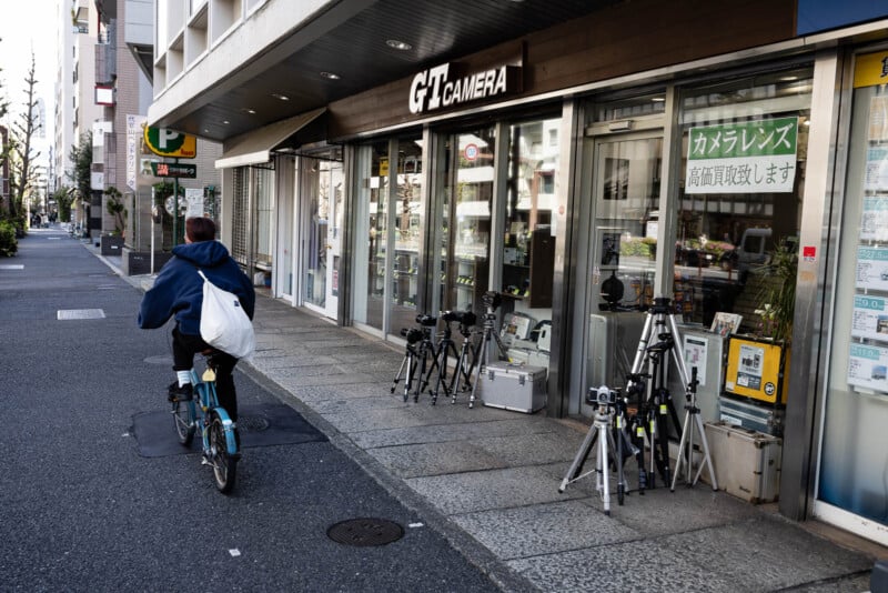 A person on a bicycle rides past a camera store with several tripods and cameras displayed on the sidewalk outside the shop. The store has large windows and signs, and the street appears quiet.