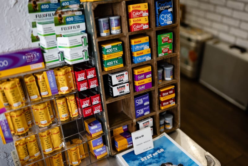 Various boxes of film rolls from brands like Kodak and Fujifilm are neatly displayed on shelves and racks in a store, alongside a price tag and part of a blue magazine or book on the counter.