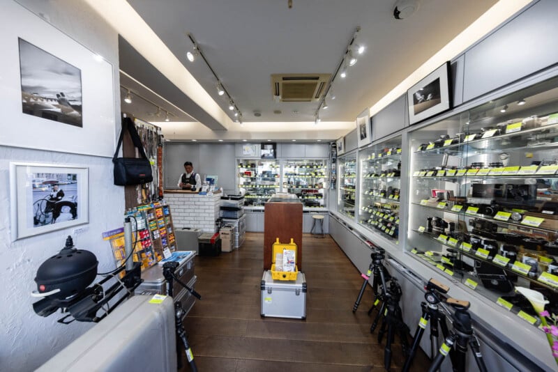 A camera store interior with glass display cases filled with cameras and lenses, tripods on the floor, framed photos on the walls, and a person standing behind the counter at the back.