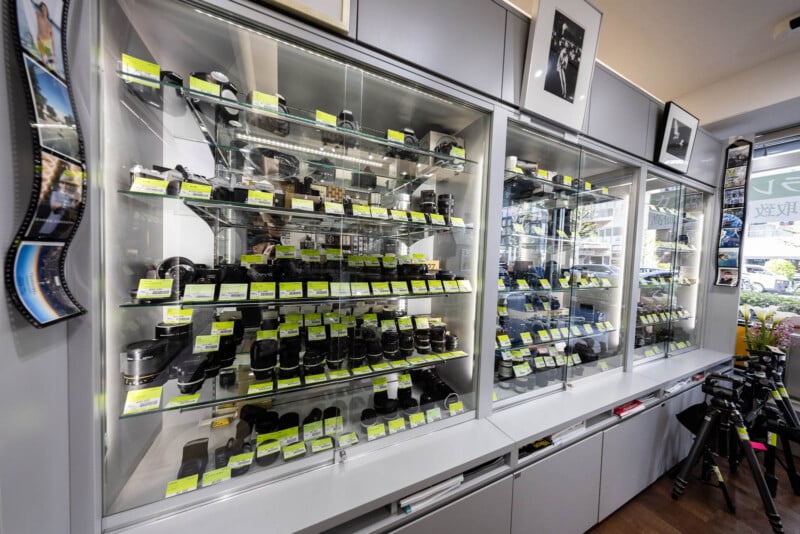 Glass display cases in a camera shop show various camera lenses and accessories, each labeled with yellow price tags. A tripod stands nearby, and black-and-white photos hang above the shelves.