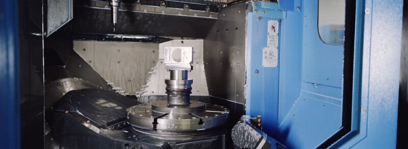 A close-up view of a CNC machine milling a metal part inside an industrial workshop, with blue panels and metallic surfaces surrounding the work area.