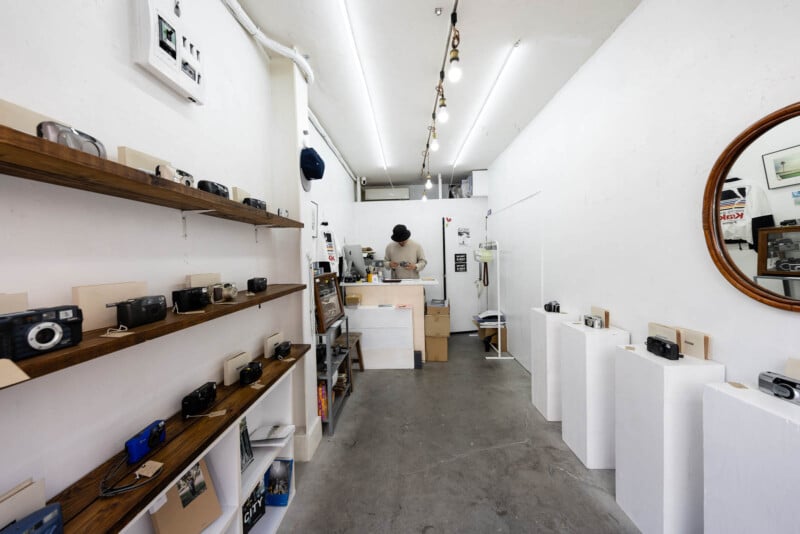 A small, minimalist camera shop with white walls, vintage cameras displayed on shelves and pedestals, and a person standing behind a counter at the back of the store.