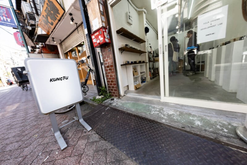 A sidewalk sign reading "KUNGFU" stands outside a shop with a glass door, through which several people are visible inside. The shop is on a narrow urban street with other small businesses.