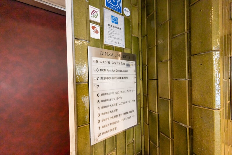 A sign listing occupants of the Ginza Church building, with floors and company names in Japanese and English, mounted on a shiny green tiled wall next to a maroon door.