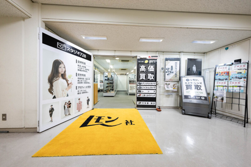 A bright indoor hallway with a yellow carpet reading "レモン社." To the left, posters show a woman and product ads. Ahead is a camera shop entrance with Japanese signs and displays for buying and selling cameras.