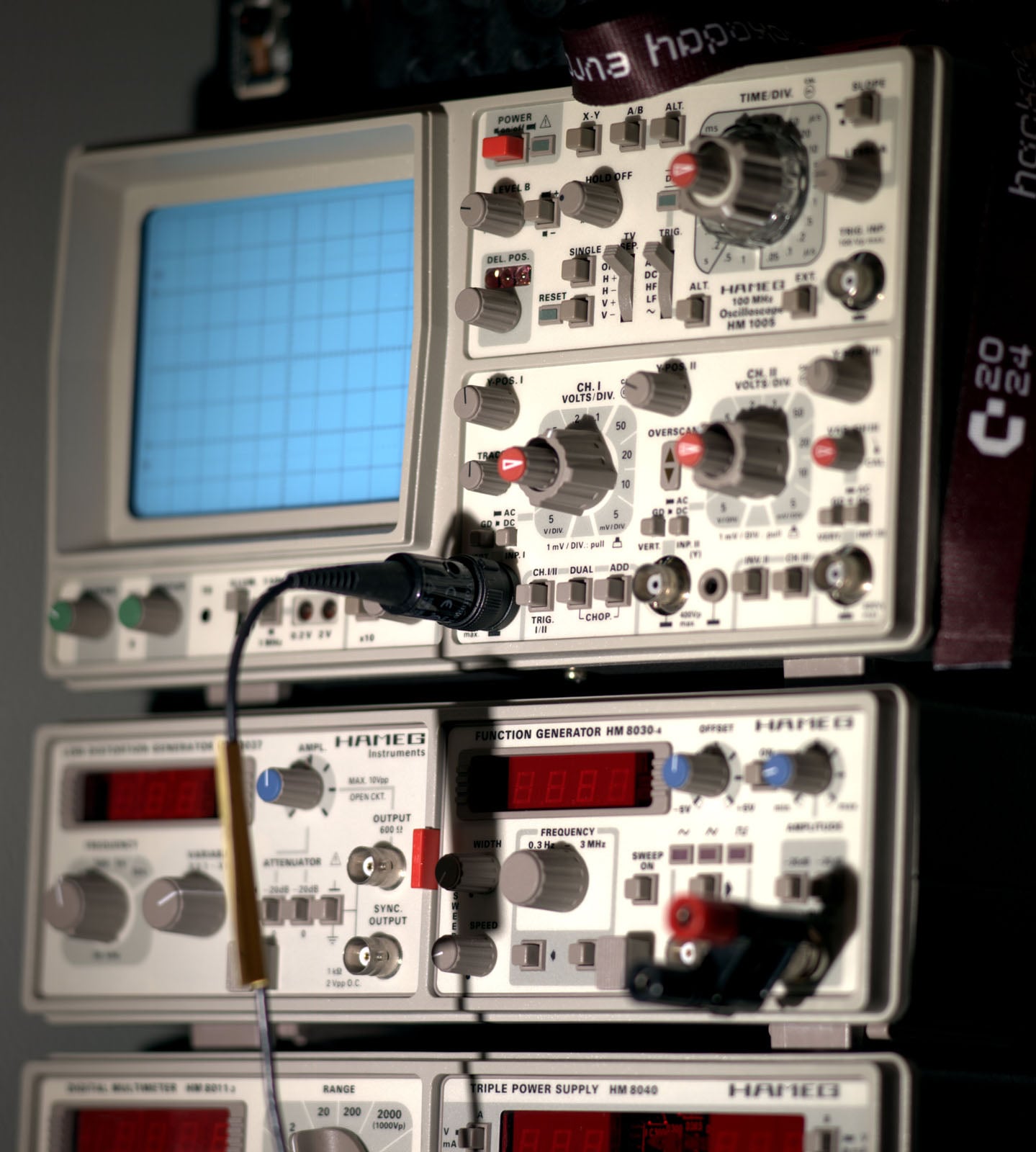 A stack of electronic lab equipment, including an oscilloscope with a blue screen, various knobs and switches, and digital function generators below, all connected with cables for testing electronic circuits.