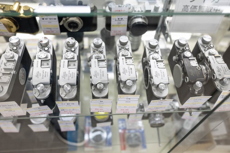 A glass display case showing several vintage Leica cameras arranged in a row, each with price tags in Japanese attached below them.
