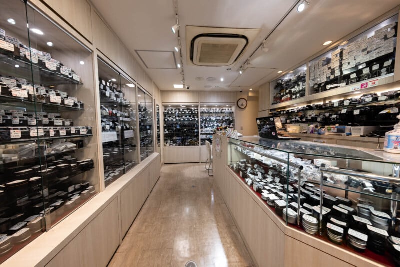A well-lit camera store with glass display cases and shelves filled with cameras, lenses, and accessories. The store has light-colored walls, a clock, and various items neatly organized throughout.