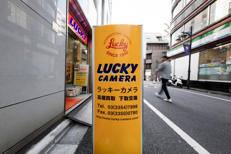 A yellow sign for "Lucky Camera" with contact details stands outside a camera store on a city street in Japan; a blurred person walks past and buildings line the background.