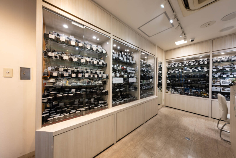A well-lit store interior with glass display cases filled with vintage and modern cameras, each labeled with small tags. The walls and cabinetry are light-colored wood, and the floor is polished marble.