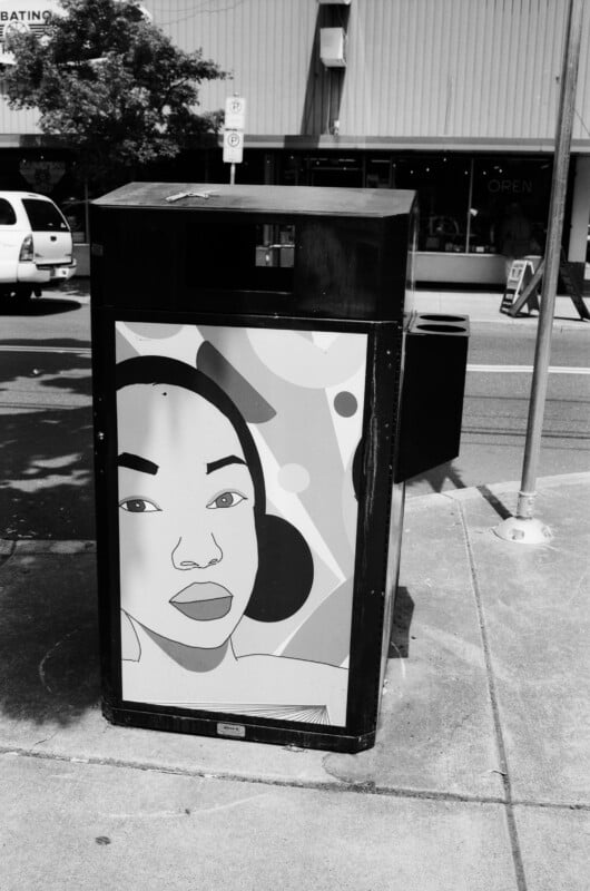 A black and white photo of a city sidewalk shows a trash bin decorated with colorful abstract art featuring a woman's face. Cars, a building, and a shop sign are visible in the background.