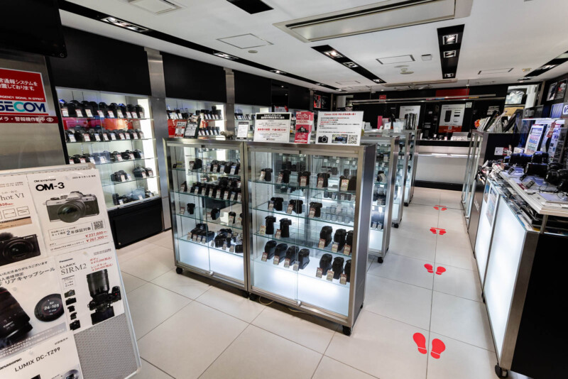 A brightly lit camera store interior with glass display cases containing various camera lenses and equipment. Signs and product information are visible, and red footprints mark a walking path on the white tiled floor.
