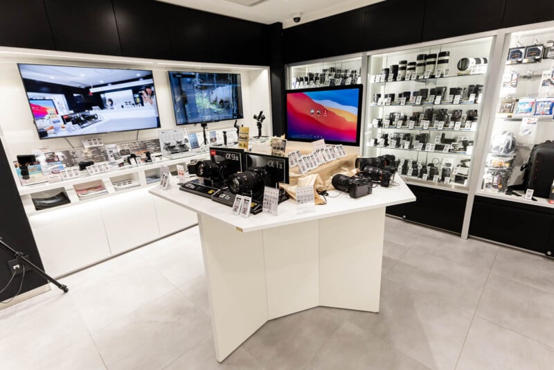 A modern camera store with digital cameras and lenses displayed on a central counter and shelves. Large screens and glass cases with camera equipment line the walls. The store is brightly lit and organized.