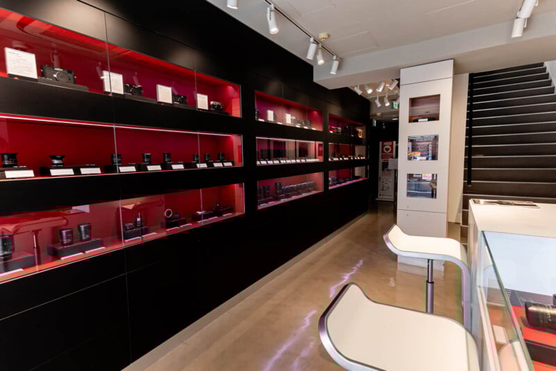 A modern camera store interior with black shelves displaying cameras and lenses against a red background, sleek white chairs at a counter, and a staircase leading upstairs. The space is brightly lit and well-organized.