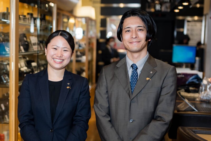 Two people in business suits smile as they stand side by side inside a well-lit store with glass display cases and shelves filled with products in the background.
