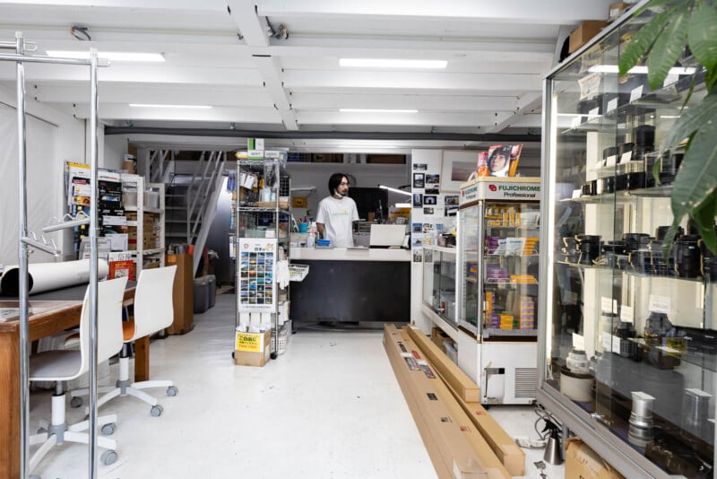 A person stands behind the counter of a camera shop, surrounded by shelves and glass cases filled with cameras, film, and photography equipment. The store has white walls, bright lighting, and an organized layout.