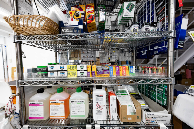 A metal shelving unit holds various boxes of analog camera film, bottles of photographic chemicals, and film accessories, organized neatly on different shelves in a store or workspace.