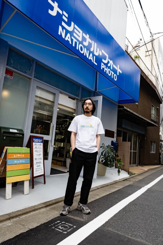 A man with glasses and a beard stands on a street in front of a blue-and-white storefront that reads “National Photo” in English and Japanese. He wears a white t-shirt, black pants, and sneakers.