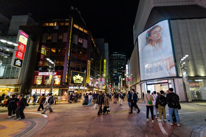 A busy street in a city at night with crowds of people crossing, bright store signs, and a large digital billboard displaying a woman's portrait on the side of a building.