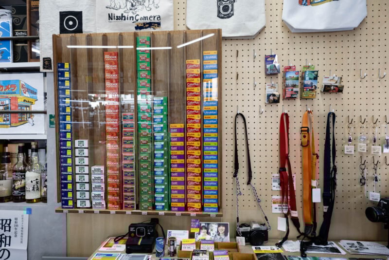 A display of colorful film rolls stacked vertically in a glass case, surrounded by vintage cameras, camera straps, postcards, and photography accessories on a pegboard wall in a camera shop.