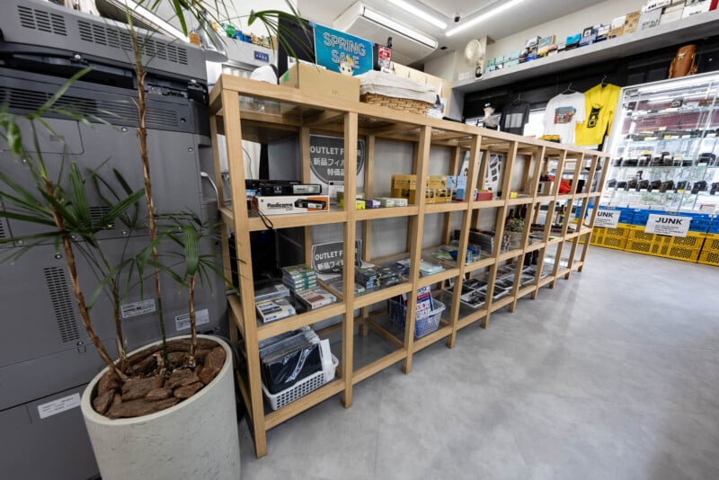 A wooden shelf displays various electronics, gadgets, and boxes in a brightly lit store with modern decor. A potted plant is on the left, and yellow crates and merchandise are visible near the window on the right.