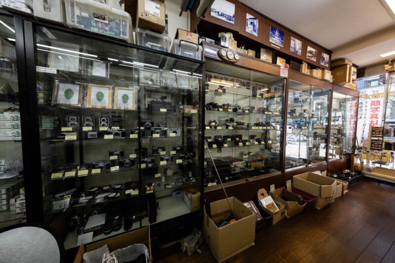 A camera shop interior with glass display cases filled with vintage cameras and equipment, boxes of items on the wooden floor, and shelves lined with photography accessories and memorabilia.