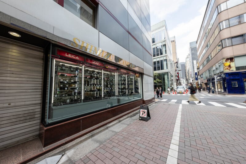 A city street corner with a camera shop called Shimizu Camera. The shop window displays various cameras. Pedestrians walk across the street; modern buildings and a crosswalk are visible.