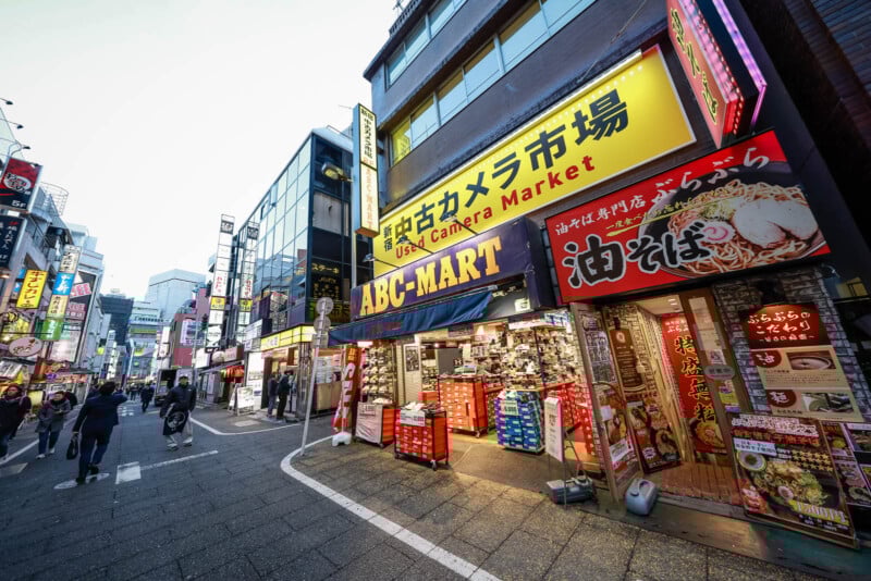 A bustling street in Japan with various storefronts, including ABC-MART and a used camera market, colorful signs in Japanese, and people walking along the sidewalk.