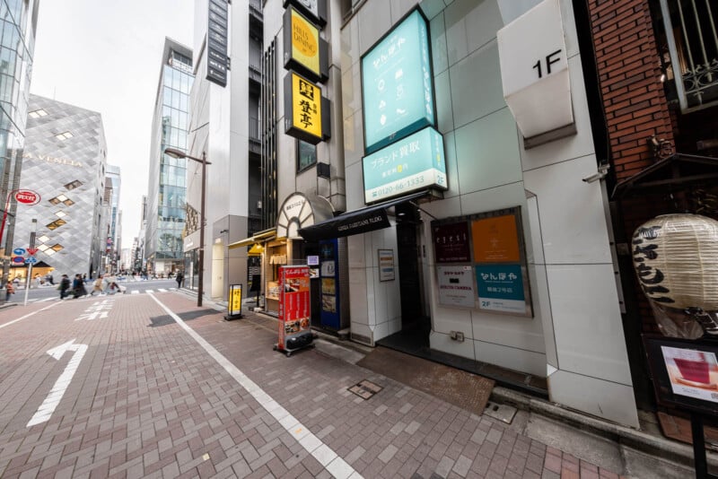 A city street in Japan with modern buildings, Japanese signs, a vending machine, and a large paper lantern outside a restaurant. The street is paved with bricks and has a few pedestrians in the distance.