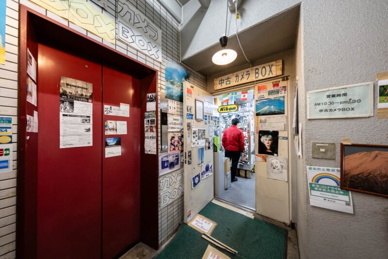 Entrance to a small Japanese camera shop with a red elevator on the left and walls covered in posters, photos, and camera ads. A person in a red jacket stands inside the brightly lit store.