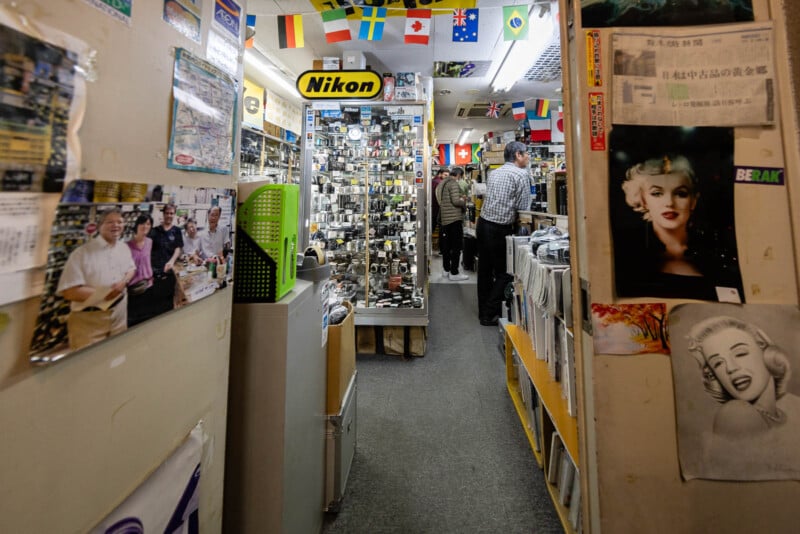 Narrow aisle in a camera store with Nikon sign, various cameras on display, international flags hanging from the ceiling, posters on the walls, and people browsing inside.