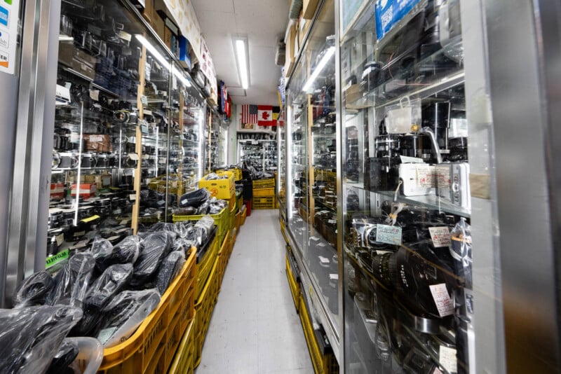 A narrow aisle in a camera shop lined with glass display cases filled with various camera lenses and equipment, with yellow bins of gear on the floor and flags hanging above.