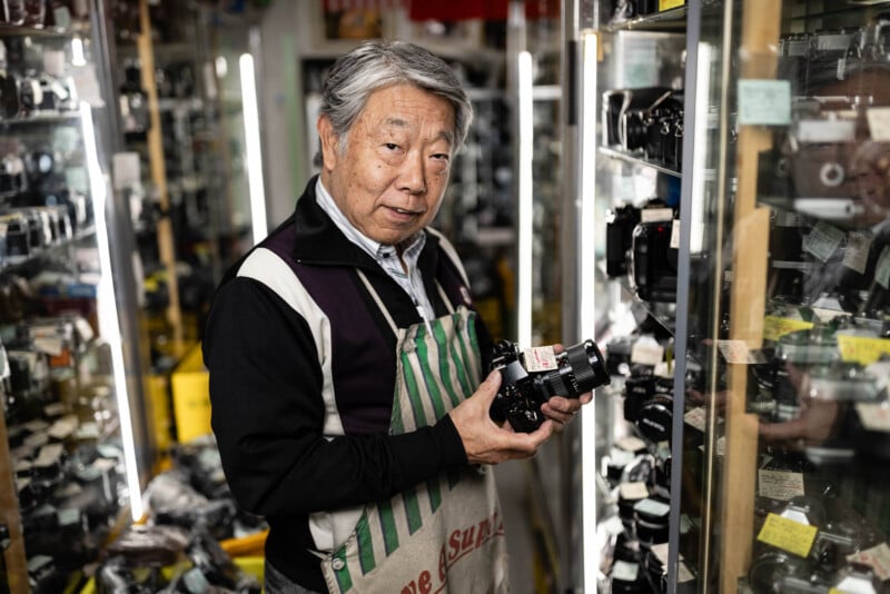 An older man wearing an apron stands in a camera shop, holding a vintage camera and looking at the camera. Shelves filled with various cameras and equipment surround him.