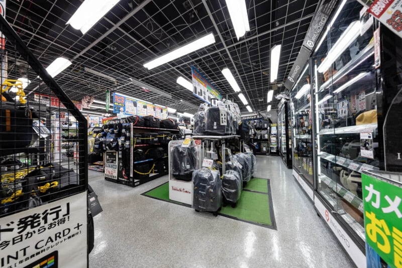 A brightly lit store interior with shelves and racks displaying backpacks and electronics. Signs with Japanese text are visible above the aisles, and glass display cases line the right side of the walkway.