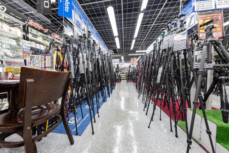 Rows of camera tripods on display in an electronics store, with bright lighting, informational signs in Japanese, and a wooden chair visible on the left side of the aisle.
