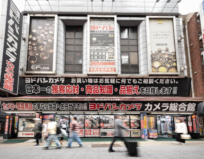 A busy street scene in front of a large Japanese electronics store, Yodobashi Camera, with people walking by. The storefront displays large banners, camera advertisements, and colorful signs in Japanese.