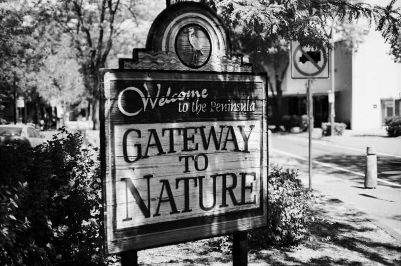 A wooden sign reads "Welcome to the Peninsula, Gateway to Nature" on a leafy sidewalk, with trees and a road visible in the background. The scene is in black and white.
