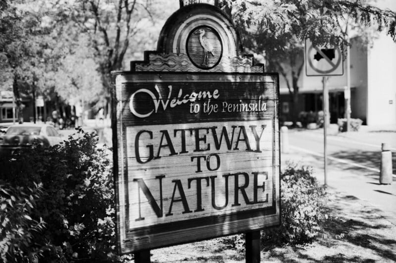 A wooden sign reads "Welcome to the Peninsula - Gateway to Nature," surrounded by trees and bushes, with a road and street signs visible in the background.